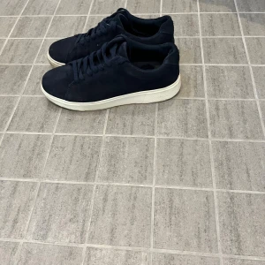 Mörkblå sneakers med vit sula - Snygga mörkblå sneakers med klassisk rund tå och platt vit sula. Skorna har snörning och är tillverkade i mocka för en mjuk och stilren känsla. Perfekt för dig som gillar enkel och clean design med en modern touch.