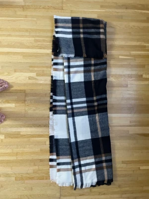 Rutig halsduk i svart, grått och beige - Stor och mjuk halsduk med rutigt mönster i svart, grått, vitt och beige. Klassisk design med fransar i kanterna, perfekt att svepa runt halsen för extra värme. Passar dig som gillar stilrena accessoarer med lite edge.