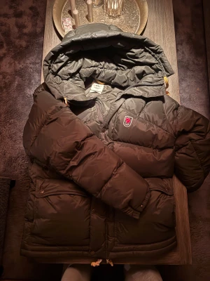 Mörkgrön pufferjacka från Fjällräven - Säljer en mörkgrön pufferjacka från Fjällräven med huva och klassisk röd logga på bröstet. Jackan har dragkedja, knappar och stora fickor framtill. Materialet är syntet och jackan är quiltad för extra värme. Perfekt för kalla dagar.