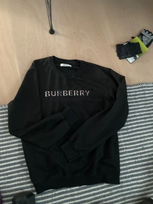 Svart sweatshirt från Burberry - Svart sweatshirt från Burberry med klassisk logga broderad på bröstet i rutigt mönster. Tröjan har rund hals, ribbade muddar och lång ärm. Perfekt för dig som gillar stilrena designer med en touch av lyx.