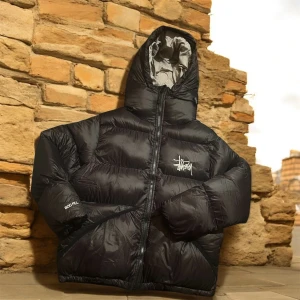 Svart pufferjacka från Stüssy - Svart pufferjacka från Stüssy med fet logga på bröstet och huva. Jackan har quiltad design, dragkedja framtill och är tillverkad i syntetmaterial. Perfekt för kalla dagar och har en clean streetwear-vibe.