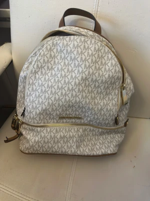 Michael Kors beige ryggsäck med MK-mönster - Snygg ryggsäck från Michael Kors i beige och ljusbrun med MK-logga över hela väskan. Väskan har flera fack med guldfärgade dragkedjor och justerbara remmar i brunt skinn. Perfekt storlek för att få plats med allt du behöver och stilren design.