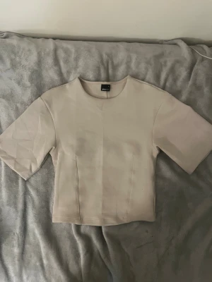 Beige croppad topp från Gina Tricot - Snygg beige croppad topp från Gina Tricot med korta ärmar och rund hals. Toppens stilrena design har diskreta sömmar framtill och baktill som ger en cool touch. Perfekt att matcha med jeans eller kjol för en clean look.