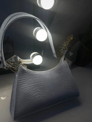 Ljusgrå axelväska i croco-imitation - Snygg ljusblå axelväska med croco-mönstrad yta och guldfärgade metalldetaljer. Väskan har en stilren, avlång form och stängs med dragkedja upptill. Perfekt accessoar för att lyfta din outfit med en trendig touch.