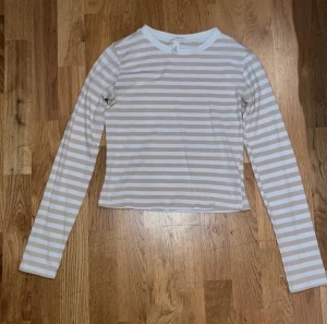 Beige och vit randig långärmad topp - Säljer en fräsch långärmad topp med beige och vita horisontella ränder. Toppen har en rund halsringning och är croppad i modellen. Perfekt för dig som gillar enkel och stilren design med en chill vibe. Har några minimala fläckar men inget som syns.