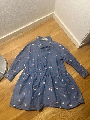 Blå blommig skjortklänning - Supersöt blå skjortklänning med små vita och rosa blommor. Klänningen har lång ärm, knappar framtill och en avslappnad passform med markerad midja. Perfekt för dig som gillar en chill och feminin stil.