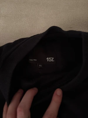 Svart sweatshirt från 157 XS - En svart sweatshirt från 157 i storlek XS. Tröjan har rund hals, långa ärmar och ribbade muddar vid ärmslut och nederkant. Enkel och clean design som funkar till jeans eller mjukisbyxor. Perfekt för dig som gillar basic och stilrent.