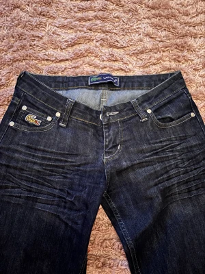 Bootcut jeans från Lacoste, mörkblå - Skit Snygga mörkblå bootcut jeans från Lacoste med klassisk krokodillogga på fickan och bak Samt låg midja. Har tyvärr växt ut dom. Jag är 170 och de passar bra på mig i längden. Dom är inte för korta. Skulle säga att de sitter bra på någon med xs. Köpte de från plick innan och de var tidigare syda till bootcut tror jag men det är inget man märker av!