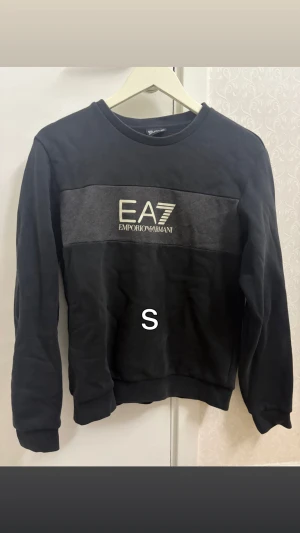 Svart sweatshirt EA7 Emporio Armani S - Svart sweatshirt från EA7 Emporio Armani med grå panel och logga framtill. Tröjan har rund hals, långa ärmar och en vit randdetalj på ryggen. Mjuk bomullskänsla och klassisk passform, perfekt för en clean och stilren look. Knappt använd.