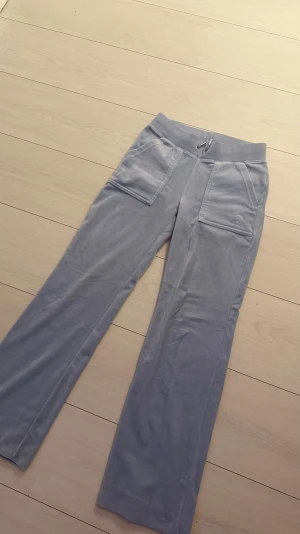 Ljusblå mjukisbyxor Juicy Couture XXS - Säljer ett par ljusblå mjukisbyxor från Juicy Couture i storlek XXS. Byxorna har bred resår med snörning i midjan, stora fickor fram och bak med metallknappar, samt broderad JC-logga på ena fickan. Materialet är mjukt och skönt, perfekt för chill. Nypris 1200kr