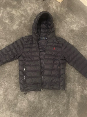 Mörkblå dunjacka från Polo Ralph Lauren - Säljer en mörkblå dunjacka från Polo Ralph Lauren med huva och dragkedja. Jackan har det klassiska röda Polo-logot på bröstet och två fickor med dragkedja. Perfekt för kalla dagar och riktigt snygg till streetwear-stilen. Extremt liten defekt på ryggen (sista bilden) som knappt syns annars nyskick. 