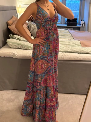 Långklänning  - En maxiklänning köpt på en marknad med färgstarkt mönster i blått, rosa och gult. Klänningen har djup v-ringning, smala axelband och markerad midja med knyt. Luftigt material och flowy passform, perfekt för sommaren.
