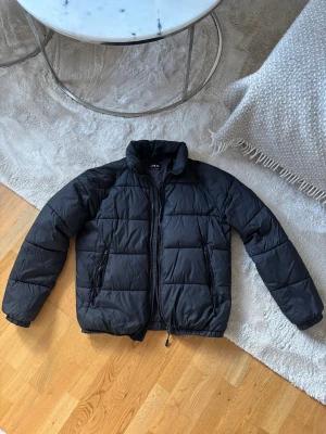 Svart pufferjacka från Zara - Svart pufferjacka från Zara med hög krage och två sidofickor med dragkedja. Jackan har ett stilrent, quiltat mönster och är tillverkad i syntetmaterial som håller dig varm under kyliga dagar. Perfekt för dig som gillar en clean och modern look.