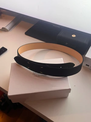 Maison Margiela svart bälte - Svart bälte från Maison Margiela i äkta italienskt skinn med rå textur och vita signaturstygn. Insidan är beige och har präglad logga. Minimalistisk och stilren design som lyfter din outfit med en lyxig touch. 