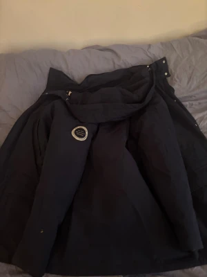 Svart parkajacka från GANT - Parkajacka från GANT i svart med grått quiltat foder och snyggt märke på ärmen. Jackan har huva, dragkedja och tryckknappar framtill. Perfekt för kalla dagar med sin långa passform och rejäla material. GANT-logga på insidan och detaljer i silver. Ny pris 3799 kr kvitto finns 