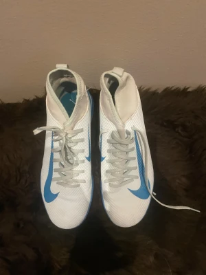 Nike vita och blå fotbollsskor - Säljer ett par vita och blå Nike fotbollsskor med snörning och platt sula. Skorna har en snygg blå swoosh på sidan och mönstrad blå yttersula med vita prickar. Ovandelen är i syntetmaterial med en modern, strömlinjeformad design och högt skaft för extra stöd.