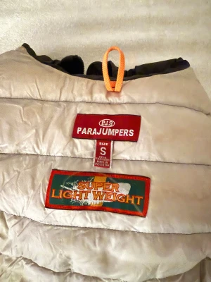 Svart lättviktsdunjacka Parajumpers S - Säljer en svart, lång och superlätt dunjacka från Parajumpers i storlek S. Jackan har quiltad design, huva och klassisk logga på ärmen. Perfekt för kyliga dagar när du vill ha en stilren look utan att frysa.