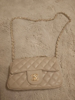 Beige quiltadl axelväska - Snygg beige axelväskal med klassiskt quiltat mönster och guldfärgad kedja. Väskan har en elegant form, ikonisk CC-logga i guld på framsidan och är tillverkad i mjukt skinn. Perfekt storlek för det viktigaste och ger en lyxig vibe till din outfit.