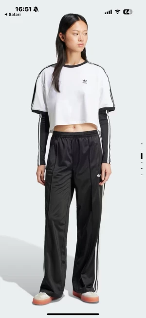 Adidas svarta wide trackpants - Svarta Adidas trackpants med klassiska vita ränder längs sidorna och broderad logga framtill. Byxorna har elastisk midja, raka och vida ben samt dragkedjefickor. Tillverkade i glansig polyester för en sportig vibe.