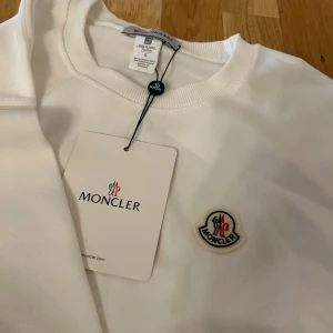 Vit sweatshirt från Moncler med logga - Vit sweatshirt från Moncler i bomull med rund hals och ribbade muddar. Moncler-logga på bröstet och stort svart M-tryck med logga på ena ärmen. Stilren och clean design, perfekt för dig som gillar exklusiva märken.