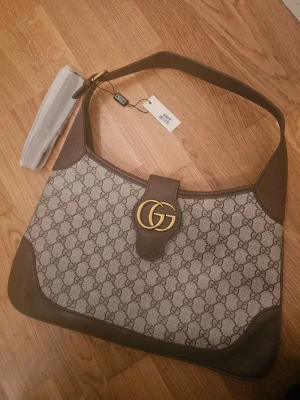 axelväska med GG-mönster - Snygg axelväska från i beige och brunt med klassiskt GG-mönster och guldfärgad logodetalj framtill. Väskan har bred axelrem, insida med fack och dragkedja, samt detaljer i skinn. Perfekt för dig som vill ha en ikonisk designerbag med plats för allt du behöver.