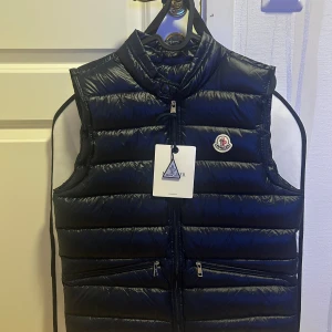Moncler GUI Väst - Snygg mörkblå dunväst från Moncler med quiltad design och klassisk logotyp på bröstet. Västen har dragkedja framtill, två praktiska fickor med dragkedja och hög krage. Perfekt för lager-på-lager och stilrena outfits.