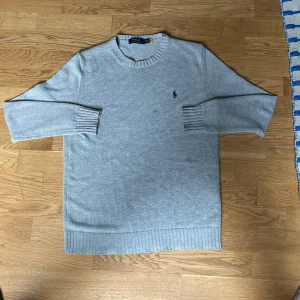 Ljusgrå stickad tröja från Polo Ralph Lauren - Säljer en ljusgrå stickad tröja från Polo Ralph Lauren med klassisk rund halsringning och broderad marinblå logga på bröstet. Tröjan har långa ärmar och ribbade muddar vid ärmslut och nederkant. Perfekt för lager-på-lager-stil.