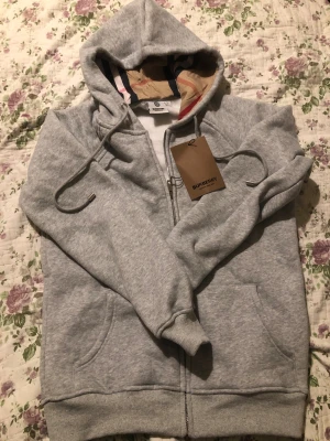 Grå hoodie från Burberry med dragkedja - Säljer en grå hoodie från Burberry i de står att stor leken är L men känns mer som en S eller lite M. Tröjan har huva med klassiskt Burberry-mönster på insidan, snörning och praktisk dragkedja framtill. Tillverkad i mjuk bomull och har två fickor fram. Perfekt för chill dagar och snygg streetstyle.