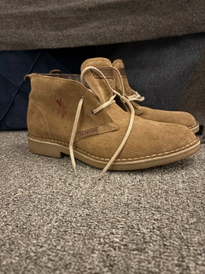 Beiga mocka kängor från Polo Ralph Lauren - Snygga beiga kängor i mocka från Polo Ralph Lauren med klassisk snörning och broderad logga på sidan. Skorna har rund tå, platt sula och detaljer i ljusbrunt. Perfekta för dig som gillar stilren och avslappnad look.