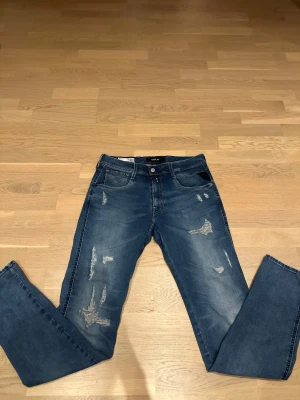 Replay Hyperflex blå jeans med slitningar - Säljer ett par Replay Hyperflex jeans i blå tvätt med coola slitningar och lagda detaljer på benen.  L34. Klassisk femficksmodell med snygga kontrastsömmar och läderpatch bak i midjan. Stretchigt material för extra komfort och stil.
