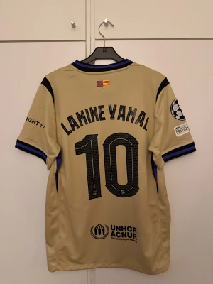 Barcelona Lamine Yamal  -  Beige FC Barcelona fotbollströja med Lamine Yamal och nummer 10 på ryggen. 