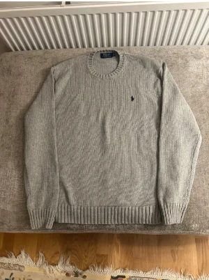 Grå stickad tröja Polo Ralph Lauren XL - Snygg grå stickad tröja från Polo Ralph Lauren i storlek XL. Klassisk rund hals och långärmad modell med ribbade muddar och nederkant. Diskret Polo-logga broderad på bröstet. Perfekt för dig som gillar stilrena och tidlösa plagg.