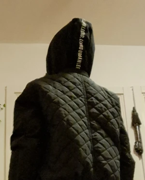 Svart quiltad bomberjacka med huva - Svart bomberjacka från Urban Legend med quiltat mönster och huva med tryckt text. Jackan har dragkedja framtill, två fickor med tryckknappar och en extra ficka med dragkedja på ärmen. Ribbad mudd vid ärmslut och nederkant för extra streetkänsla.