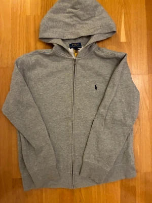Grå hoodie från Polo Ralph Lauren - Snygg grå hoodie från Polo Ralph Lauren med dragkedja och huva. Tröjan har lång ärm och klassisk Polo-logga broderad i marinblått på bröstet. Tillverkad i mjuk bomull, perfekt för chill dagar. Storlek L (14-16).