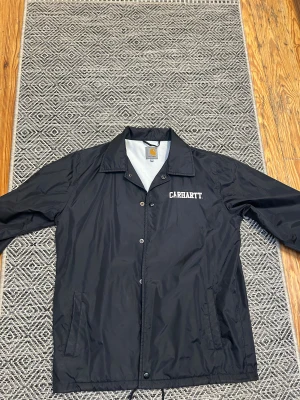 Svart Carhartt vindjacka S - Svart vindjacka från Carhartt i storlek S. Jackan har klassisk krage, tryckknappar framtill, två sidofickor och vit Carhartt-logga på bröstet. Materialet är lätt och syntetiskt, med snörning nedtill för extra passform. Perfekt för dig som gillar streetwear.