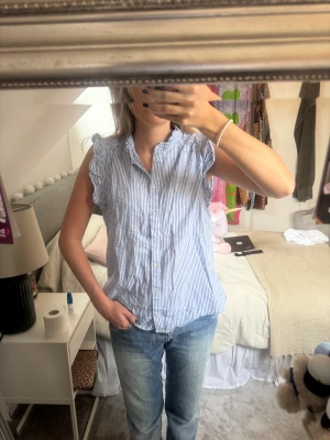 Blårandig ärmlös blus från H&M - Säljer en så söt blårandig skjorta/blus från H&M. Blusen är ärmlös med volangdetaljer vid axlarna och kragen samt har den knappar framtill. Perfekt på sommaren!