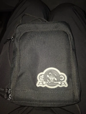 Svart axelväska med Sofialäktaren-märke - Säljer en svart axelväska med Sofialäktaren-märke framtill. Väskan har en stilren, rektangulär form och är tillverkad i slitstark polyester. Utrustad med dragkedja och justerbar axelrem, perfekt för att bära dina viktigaste grejer.