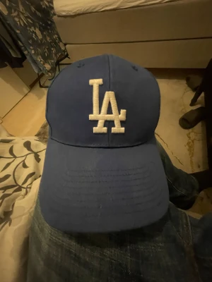 Blå LA Dodgers keps från 47 Brand - Säljer en blå LA Dodgers keps från 47 Brand med vit broderad LA-logga framtill och 47-märke på sidan. Kepsen har böjd skärm i grått och klassisk passform. Perfekt för dig som gillar sportig stil och vill representera Los Angeles Dodgers.