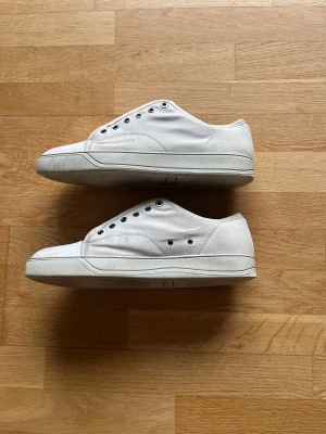 Vita sneakers från Lanvin  - Säljer ett par klassiska vita sneakers från Lanvin i Manchester  med rund tå och platt sula. Skorna har metallfärgade öljetter för snörning och en stilren design. Perfekta för dig som gillar minimalistisk och clean stil.