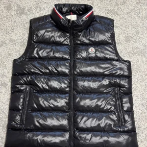 Moncler Väst svart  - Svart, glansig dunväst från Moncler med quiltad design och hög krage. Västen har dragkedja framtill, två fickor med dragkedja och klassisk Moncler-logga på bröstet. Insidan har en unik serietidningsetikett. Perfekt för lager-på-lager-stil. Storlek 9 passar mer till L eller XL  jag är xxl och den sitter tight när jag drar igen den.