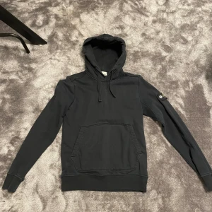 Svart hoodie från Stone Island - Svart hoodie från Stone Island med klassisk patch på vänster ärm. Tröjan har huva med snörning, stor magficka och ribbade muddar. Tillverkad i mjuk bomull, perfekt för en chill och stilren look.