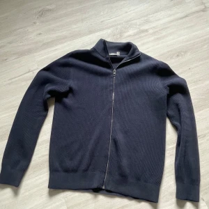 Mörkblå stickad kofta från Kappahl - Mörk, stilren stickad kofta från Kappahl i storlek S. Koftan har hel dragkedja framtill, ribbade muddar och hög krage. Perfekt för lager-på-lager och enkel att matcha med olika outfits.