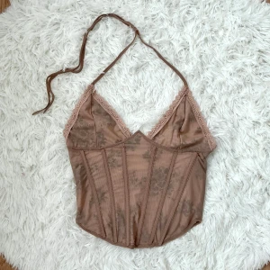 Brun blommig korsett från Karynnna - Supersnygg brun korsett från Karynnna i mesh med blommigt mönster och spetsdetaljer längs v-ringningen. Storlek S, byst 82-86cm, midjan 64-68cm.