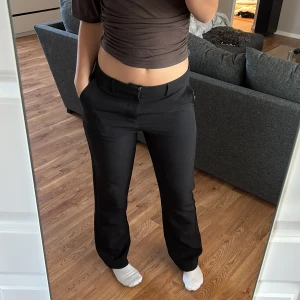 Svarta kostymbyxor med raka ben - Svarta kostymbyxor med klassisk rak passform och midja med hällor för bälte. Byxorna är högmidjade men går att vika ner om man vill det lika fint det också. Byxorna har diskreta fickor fram och bak, stängs med dragkedja och knapp. Perfekta för en clean och stilren look. Materialet känns mjukt och följsamt.