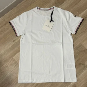 Vit Mon*ler t-shirt med logga - Vit t-shirt från Mon*ler med klassisk rund hals och korta ärmar. Slim fit-modell med Mon*ler-logga broderad på vänster ärm. Röd, vit och marinblå randig detalj på ärmslut och insidan av halsen. Tillverkad i mjuk bomull för skön känsla.