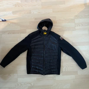 Parajumpers Nolan Mesh Hooded Hybrid Black - Svart hybridjacka från Parajumpers med huva, quiltad framsida och rygg samt stickade ärmar. Säljs då den aldrig kommer till användning. Använd max 3 gånger. Storlek XL men passar L.