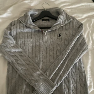 Grå kabelstickad tröja Polo Ralph Lauren - Snygg grå kabelstickad tröja från Polo Ralph Lauren med halv dragkedja och hög krage. Klassisk logga broderad på bröstet. Perfekt för dig som gillar en stilren och tidlös look. Långärmad och i mjukt stickat material.