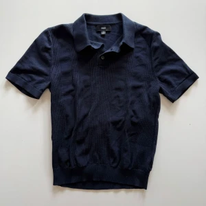 Mörkblå pikétröja från Next - Snygg mörkblå pikétröja från Next med klassisk krage och två knappar. Tröjan har korta ärmar och ribbade muddar samt diskreta vertikala strukturränder framtill. Perfekt för en stilren och avslappnad look.