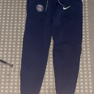 Mörkblå PSG x Nike mjukisbyxor XS - Mörkblå mjukisbyxor från Nike i samarbete med PSG. Byxorna har snörning i midjan, PSG-logga och Nike Swoosh framtill. Tillverkade i mjukt material, perfekta för chill eller sport. Snygg och sportig look med smal passform. Nypris 1300