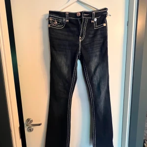True Religion jeans med vita sömmar - Mörkblå bootcut-midwaist jeans från True Religion med tydliga vita kontrastsömmar och sjukt snygga  bakfickor med broderad logga. Snygga detaljer med kopparfärgade knappar på bakfickorna. Limited edition, finns inte att köpa längre🥰